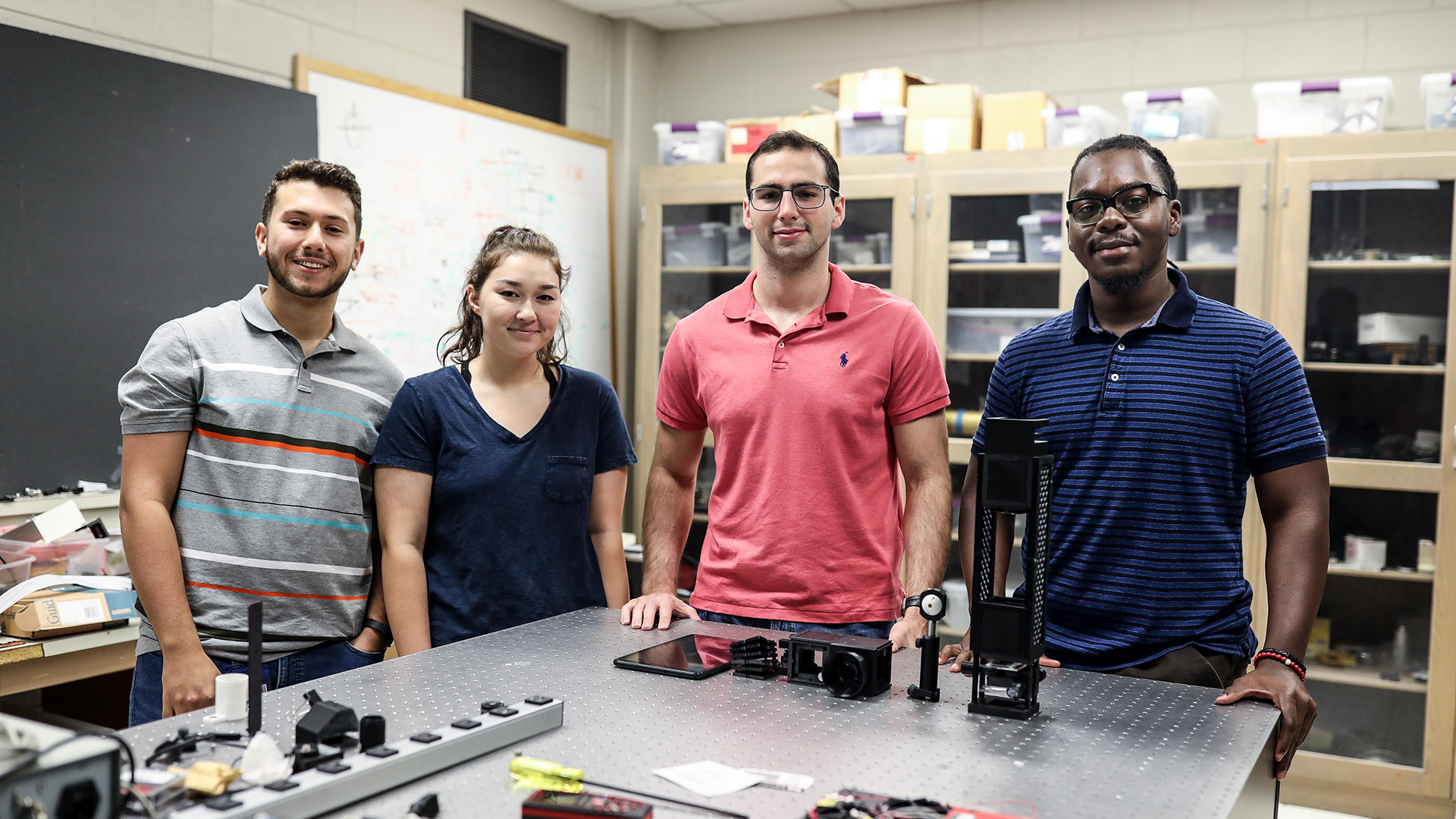 Four smiling interns (three males, one female) standing behind lab bench, looking at camera.