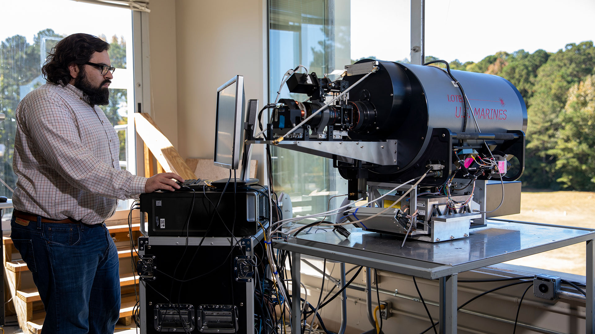 Male researcher with beard and glasses stand near the large round metal Lidar device while typing on a computer.