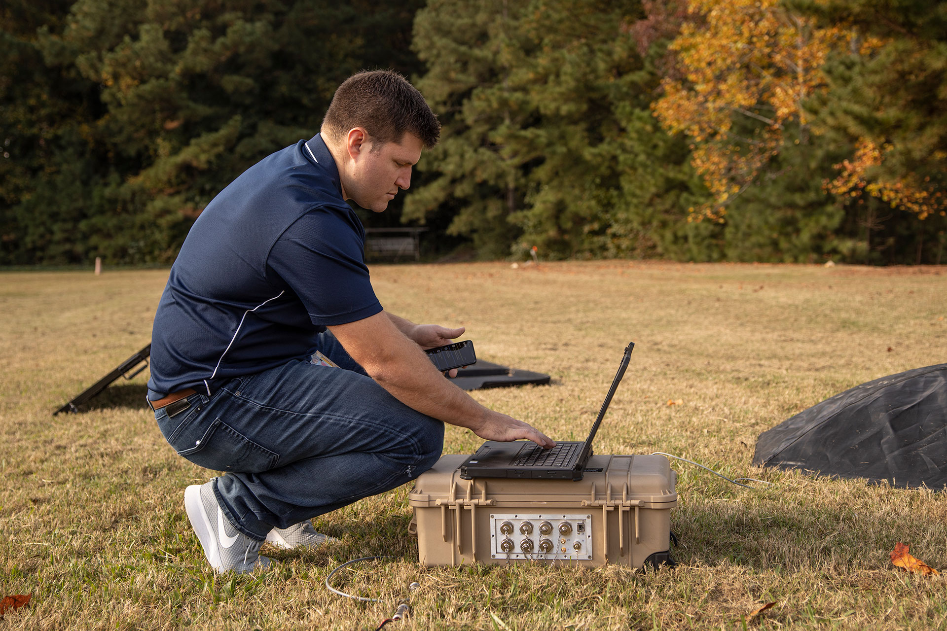Researcher outdoors squatting while typing on laptop computer that is sitting on plastic case on the ground.