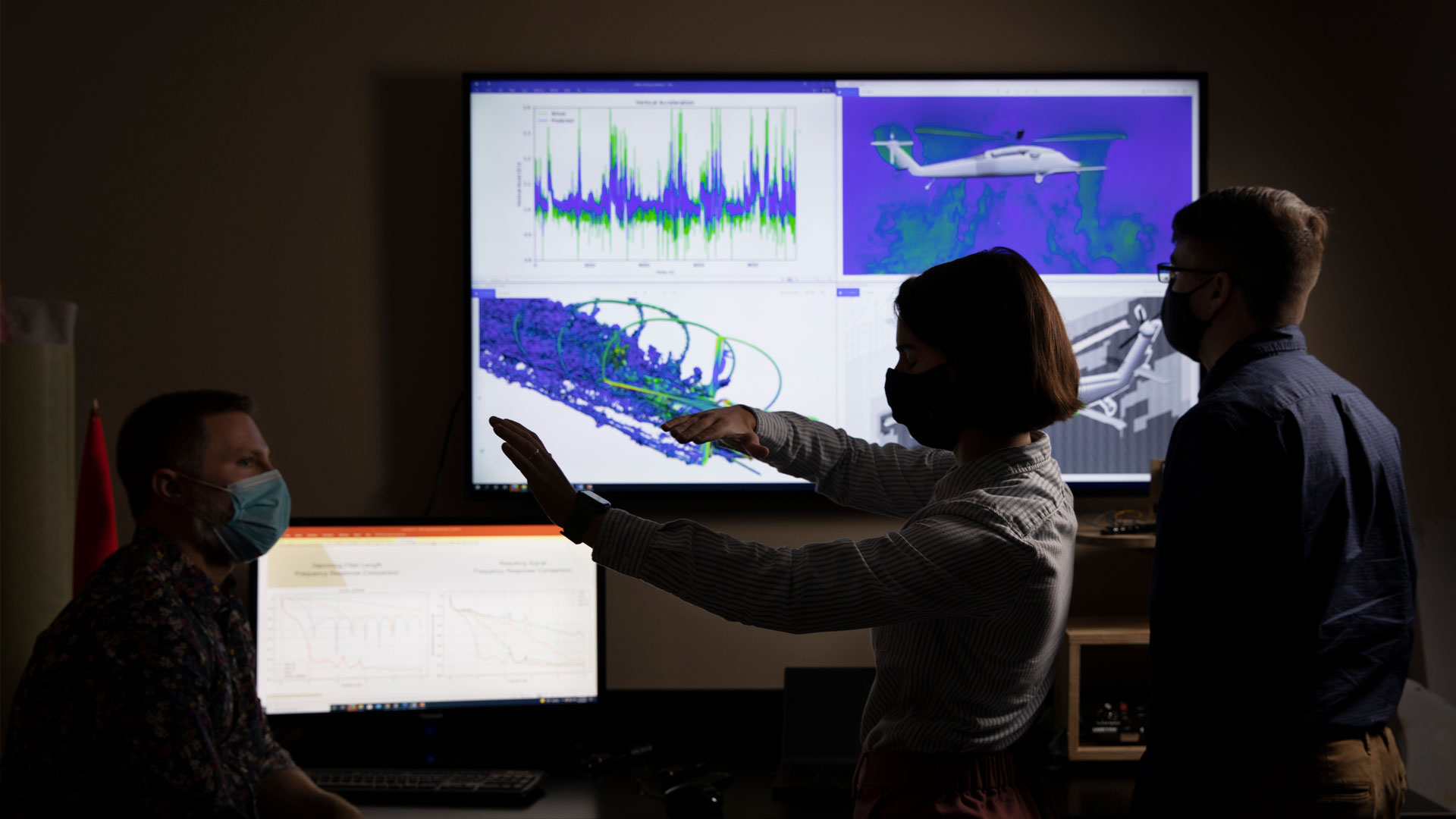 Two male and one female researcher in dark room with large video display showing brightly colored computer simulation of air flow around helicopter 