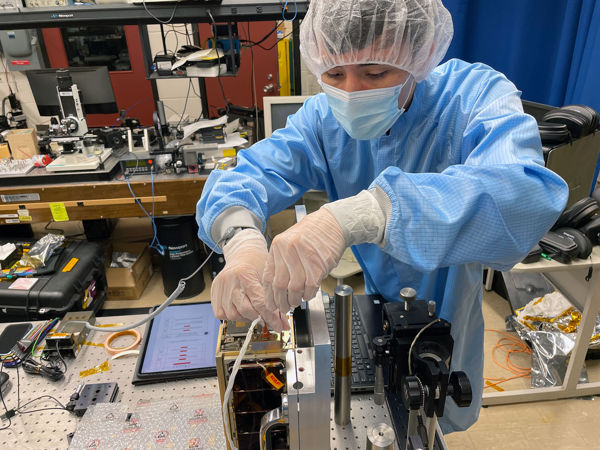 Research dressed in gown, mask, hairnet, and gloves works on device in lab.