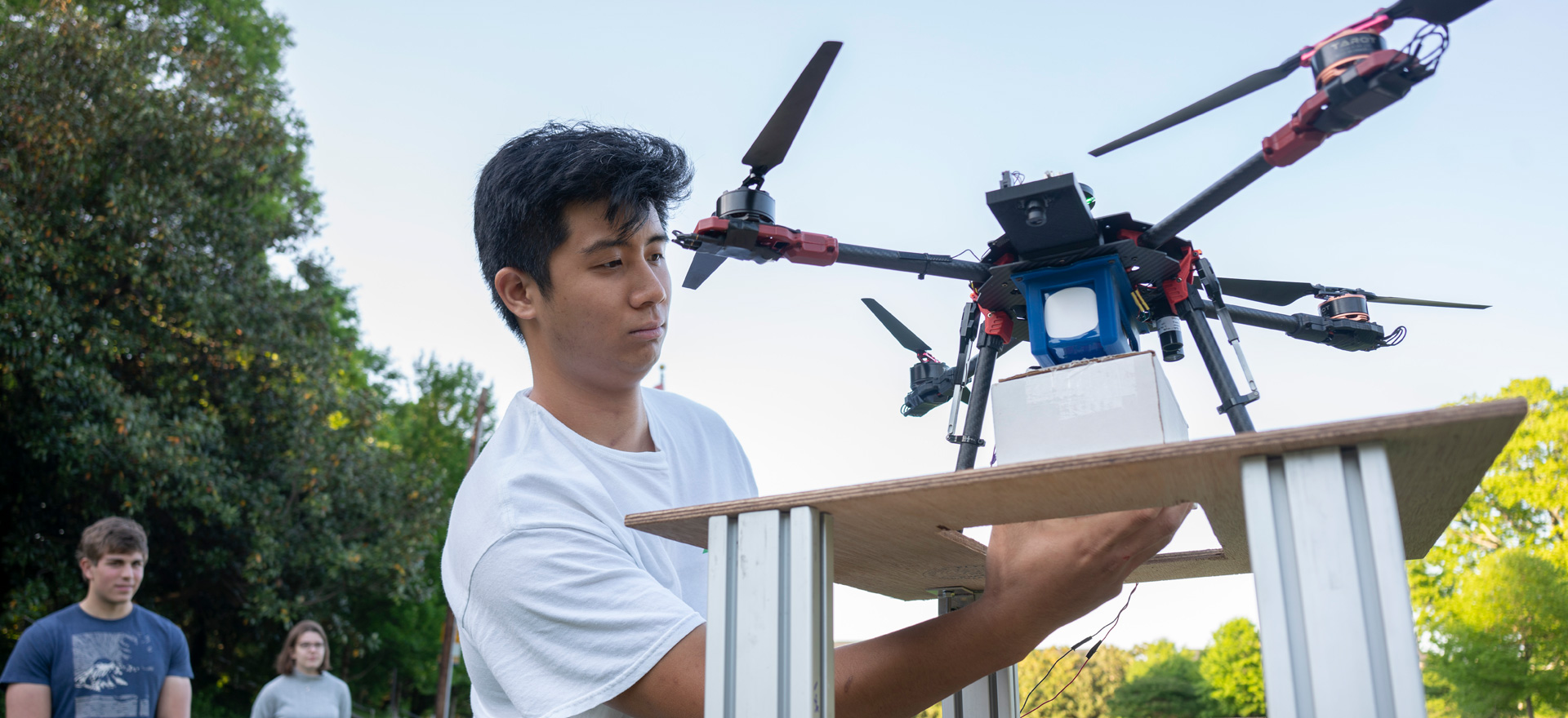Student makes adjustments to drone resting on small landing pad, outdoors in testing field.