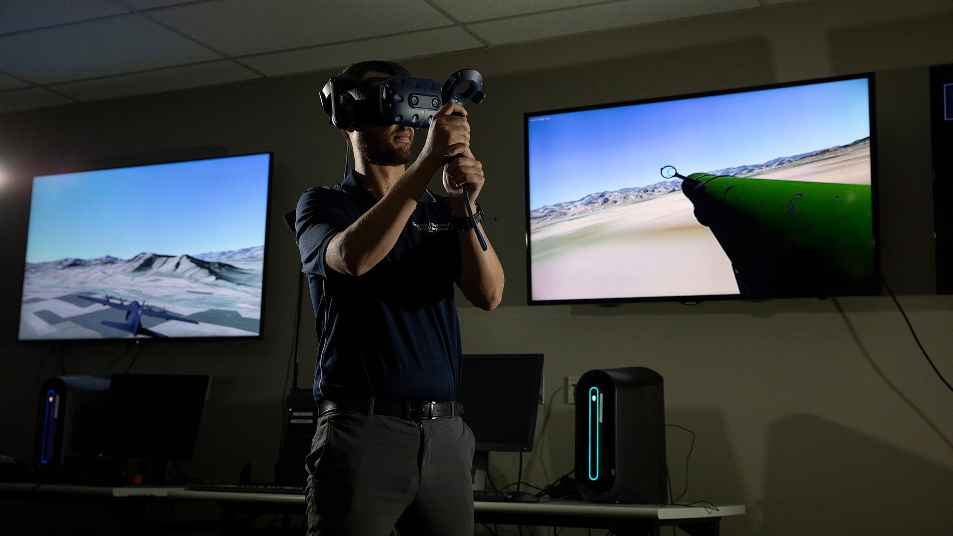 Male researcher standing in front of two large video monitors, wearing VR goggles and holding a control device in front of him