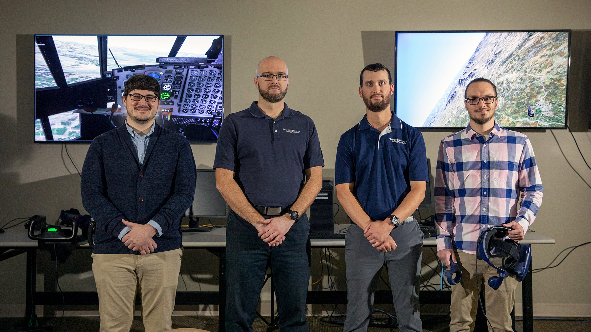 Group of 4 male researchers standing in a row posed for photo, background is FITRE setup with table and two large video monitors on wall