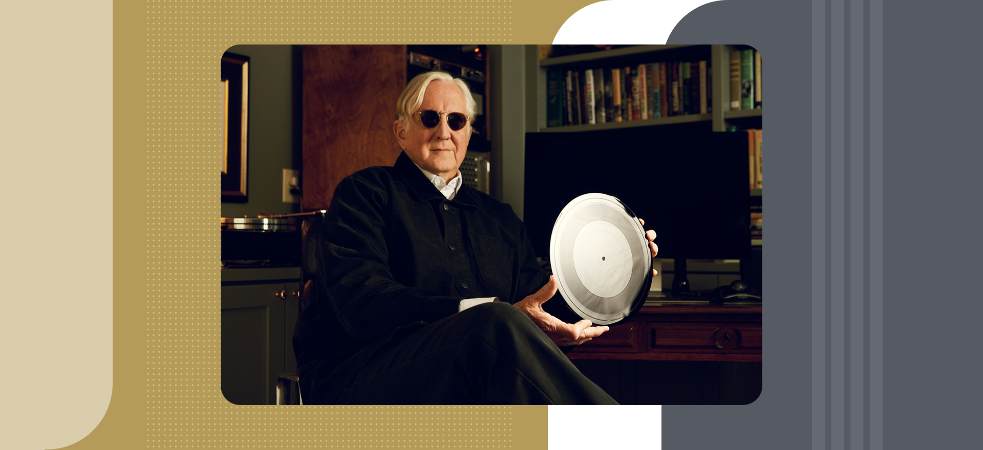 Older man wearing sunglasses, holding large silver disc, sitting in chair in dark office