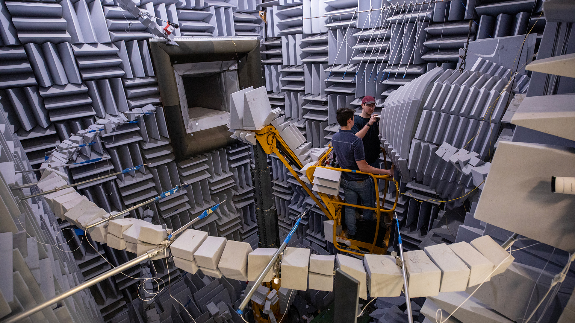 Wide view of two male researchers inside a large acoustic chamber examine a nozzle testing device.