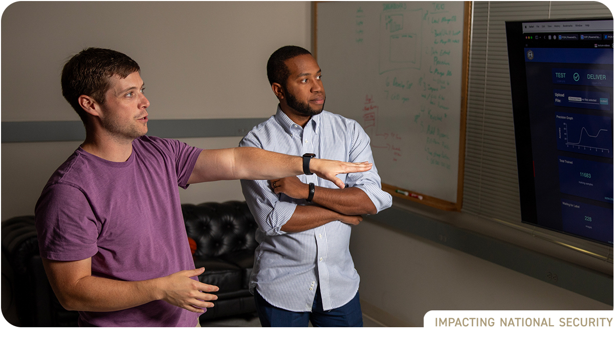 Two young male researchers stand in front of a large monitor displaying data