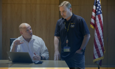 Erik is sitting at a table while talking to JD Fassett, who is standing.