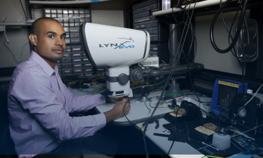 Mike Ruiz sits at a desk with a eyepiece-less microscope and various technical equipment. (Photo credit: Sean McNeil)
