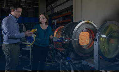 Two researchers stand near a large metal instrument with center glowing red hot