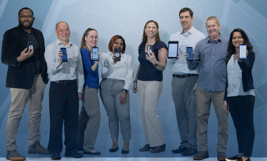 Group of eight researchers, four men and four women, dressed in street clothing, standing next to one another, holding up smartphones featuring images of app.