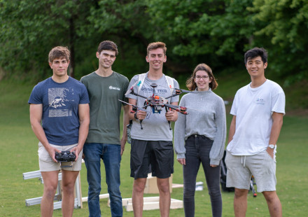 Five college students (4 males, 1 female) standing together outdoors, in a row.