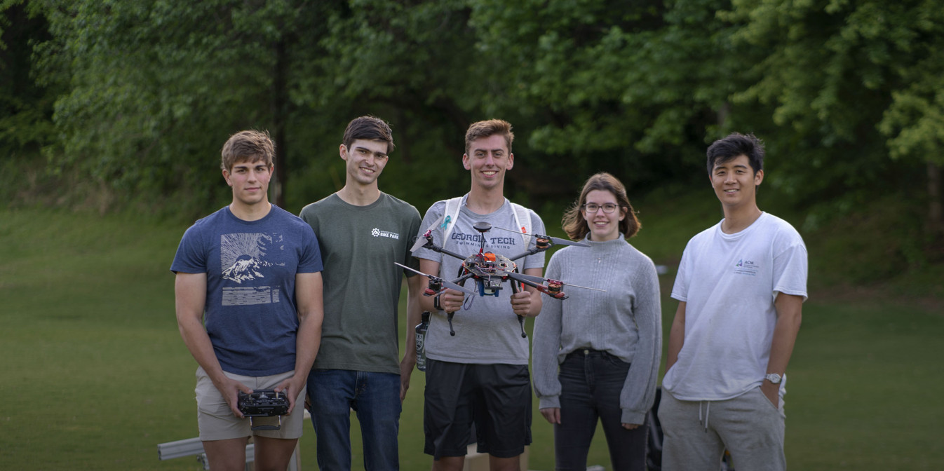 Five students standing in a row in a field smiling and looking at camera. Student in center is holding a drone.